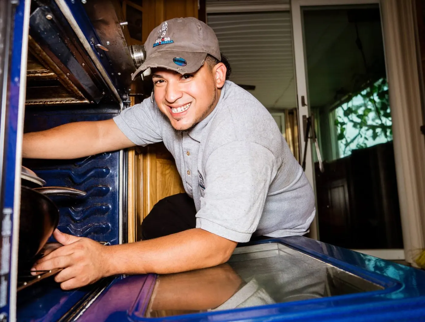 Smiling Doctor Maintenance technician in a company cap and gray polo reaching into an open oven on a Baltimore-area service call
