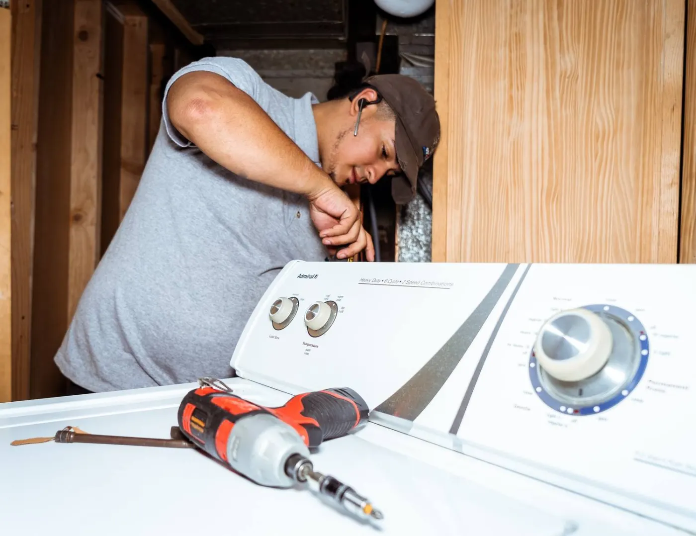 Doctor Maintenance technician reaching behind the control console of a top-load washer in a basement laundry area, an impact driver resting on the lid